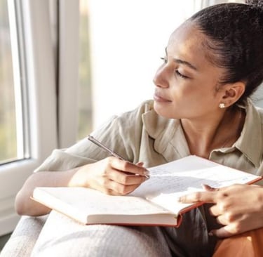 Thoughtful young woman journaling in a notebook by a window with natural light.