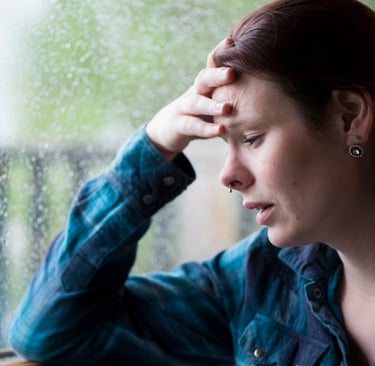 A stressed woman rests her head on her hand near a rainy window, depicting anxiety and mental health struggles.