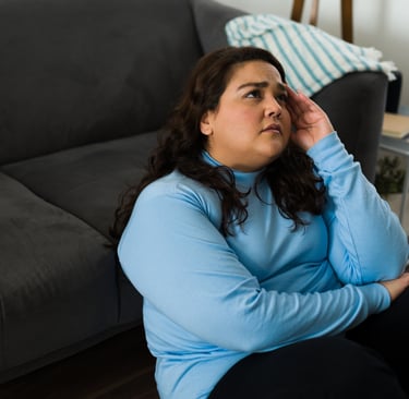 A stressed woman sitting on the floor leaning against a gray sofa, touching her head due to a headache.