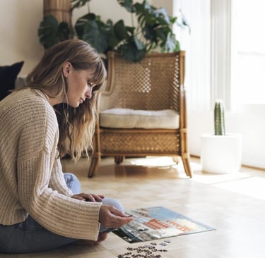 A woman sits on a wooden floor in a sunny living room assembling a jigsaw puzzle.
