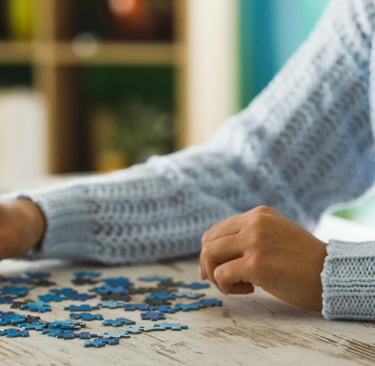 A woman in a blue sweater assembles a blue jigsaw puzzle on a rustic wooden table.