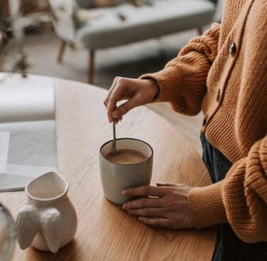 A person in a brown knit sweater stirs a cup of coffee on a wooden table.