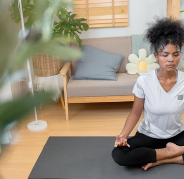 A young woman practicing yoga meditation in a lotus pose on a mat in her bright living room.