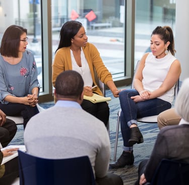 a group of people sitting in a circle with a woman in a white shirt
