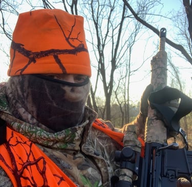 A hunter wearing a blaze orange safety vest and hat while holding a camouflaged rifle in a winter forest.