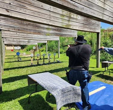 A cowboy action shooter aims a shotgun at steel targets on an outdoor shooting range.