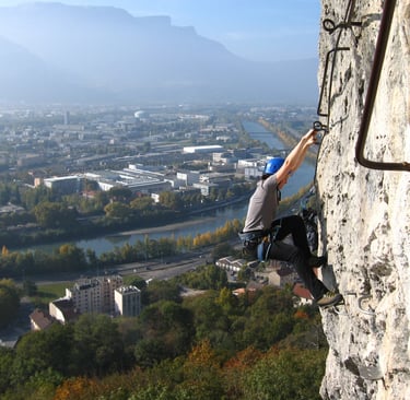 A mountain climber climbs a steep via ferrata route overlooking a scenic city valley and river.