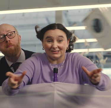 Woman in a lilac cardigan reaches forward as papers fly; man in glasses stands behind her.