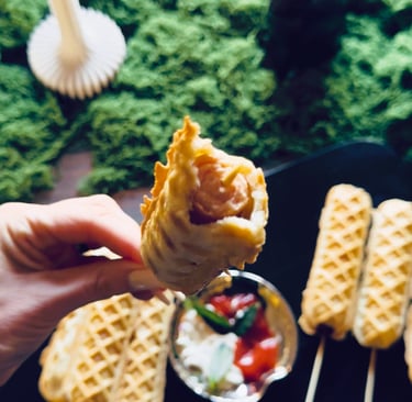 Hand holding a bitten waffle corn dog on a stick over a plate with dip and more corn dogs.