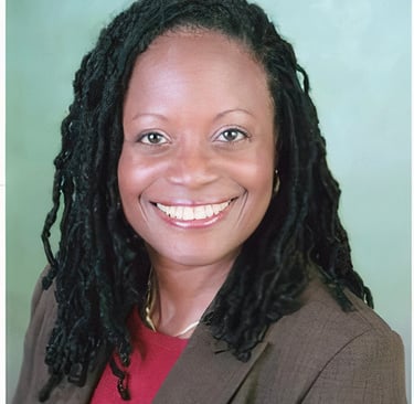 Professional portrait of a smiling woman with long dreadlocks wearing a brown blazer.