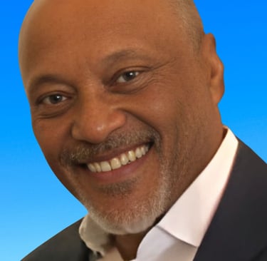 A professional portrait of a smiling bald man with a graying goatee wearing a blazer against a blue background.
