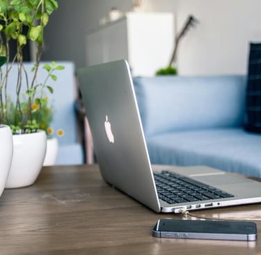 A silver MacBook laptop and iPhone on a wooden coffee table in a modern living room with green plants.