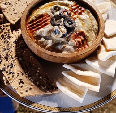 Wooden bowl of creamy hummus dip topped with olives and paprika, served with pita bread and crackers.