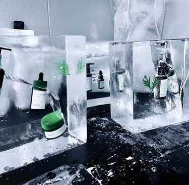 Skincare products, including a green serum bottle and jar, frozen inside clear ice blocks, with white flakes on a dark