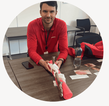 A person sits at a table holding a leg training model wrapped in a bandage, demonstrating first aid