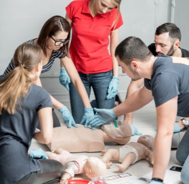 a group of people around a first aid training manikin learning CPR with an instructor