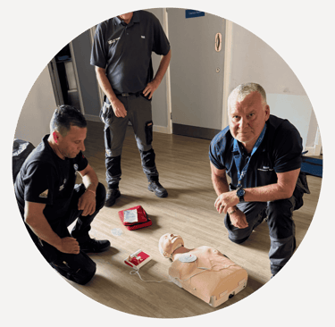 a group of men around a training manikin have attached and are using an AED.