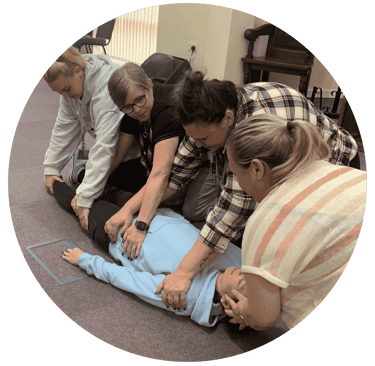 Four adults kneel around a person lying on the floor during a first aid training session