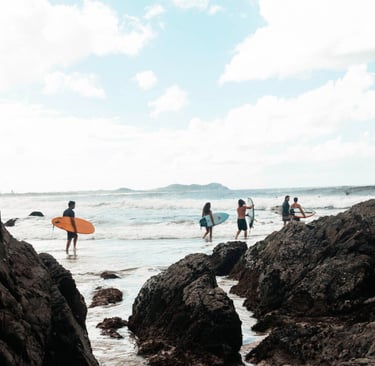 a group of surfing people walking on rocks near the ocean. Byron Bay