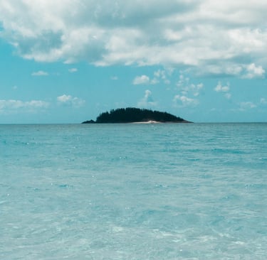 Tropical island with lush trees surrounded by clear turquoise ocean water under a cloudy blue sky.