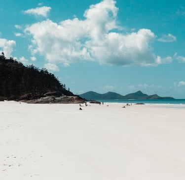 Pristine white sand beach with tourists, turquoise ocean water, and lush tropical hills under a blue sky.