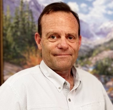 A man with dark hair, wearing a light-colored shirt. The background is a blurred landscape.
