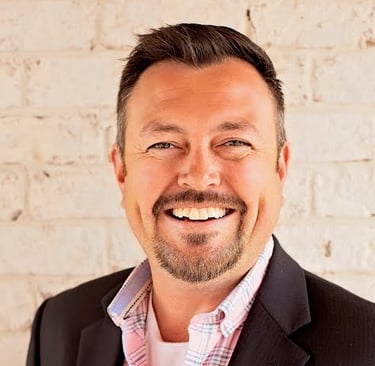 Picture of a man with brown hair and goatee, smiling. He's wearing a bright shirt and a dark suit.