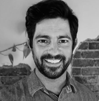 a black and white picture of a man with dark hair and beard. He is standing in front of a wall.