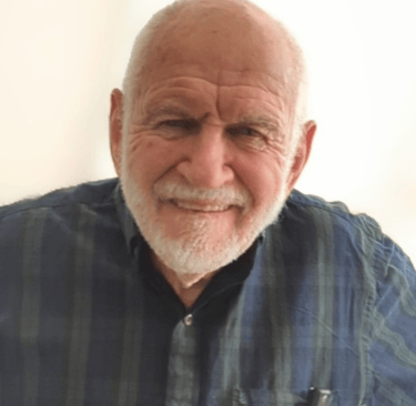 A white with a short trimmer white beard, wearing a dark patterned shirt.