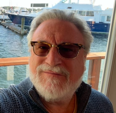 a man with a beard and sunglasses on a pier with water and boats behind him