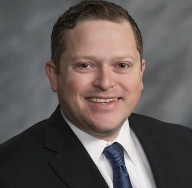 Professional headshot of a smiling man in a black suit and blue tie against a grey background.