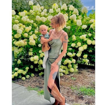 Jade in a green dress and boots holding baby Nova by flow bush, representing maternal instinct and caregiving