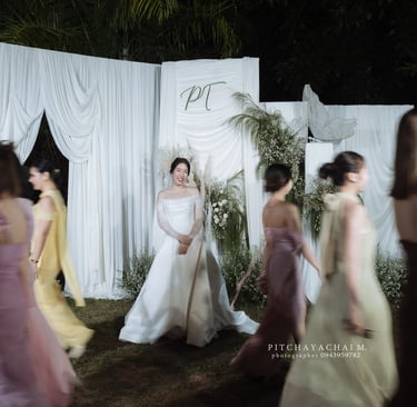 เจ้าสาวยืนกลางฉากงานแต่งโทนขาว ขณะเพื่อนเจ้าสาวเดินผ่านสร้างเอฟเฟกต์เบลอ ภาพงานแต่ง Outdoor ตอนกลางคืน