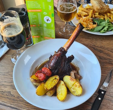 Braised lamb shank served with roasted potatoes, cherry tomatoes, and craft beer at a restaurant table.