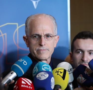 Randy Bennett speaking to reporters surrounded by microphones at a press 