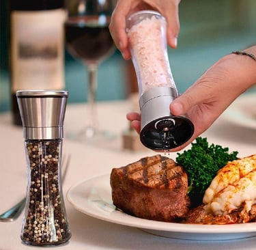 Hand using a stainless steel salt grinder over a gourmet steak and lobster dinner.