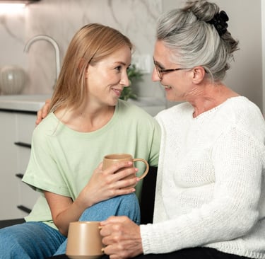 A smiling senior mother and adult daughter talking while holding coffee mugs on a sofa at home.