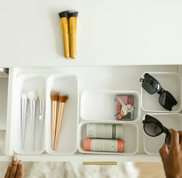 A person organizes makeup brushes and sunglasses in white plastic drawer dividers on a vanity.