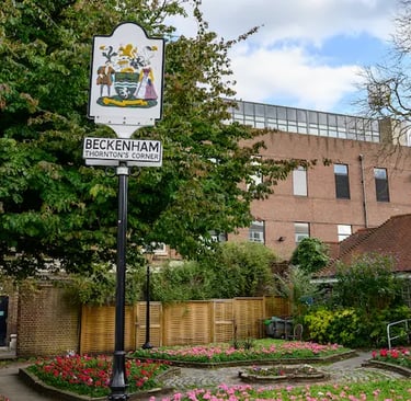 Beckenham Thornton's Corner sign standing in a public garden with flower beds and brick buildings.