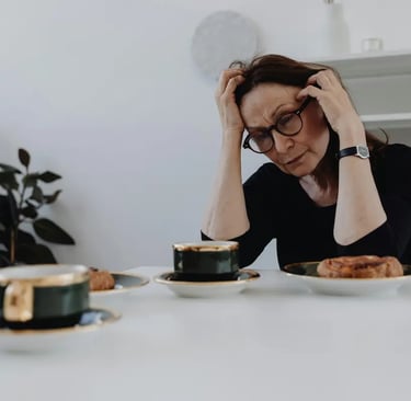 Stressed mature woman holding her head in frustration while sitting at a table with coffee and pastries.