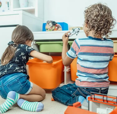 Two young children playing with toys and organized plastic storage bins in a bright playroom.