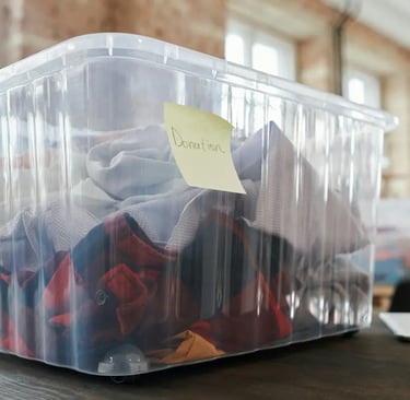 A clear plastic storage bin filled with folded clothes for donation labeled with a sticky note.