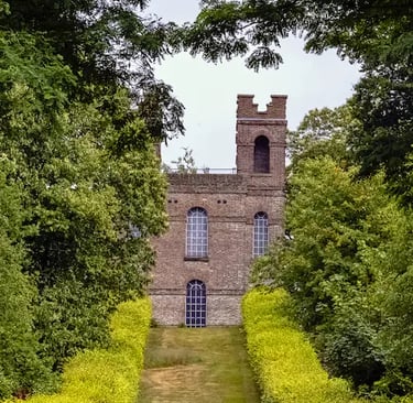 A historic brick folly castle with arched windows framed by lush green trees and a grassy pathway.