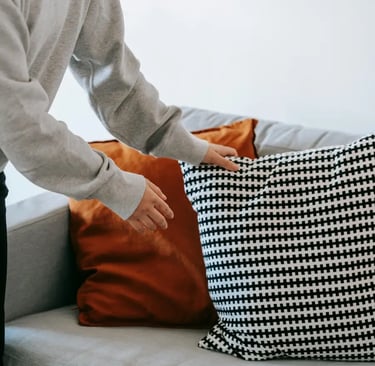 A person in a gray hoodie arranging patterned and solid orange throw pillows on a modern gray sofa.