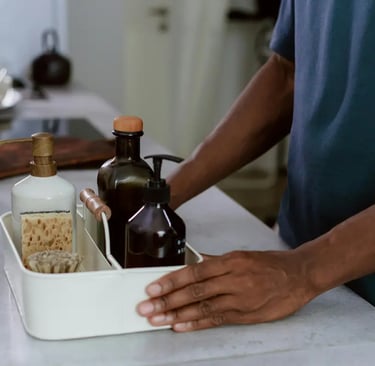 A person organizing reusable soap dispensers and cleaning tools in a white metal caddy.