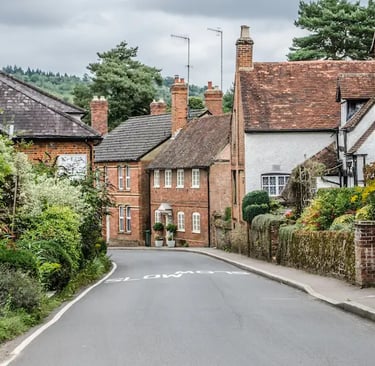 A narrow road winding through a charming English village with historic brick cottages and lush greenery.