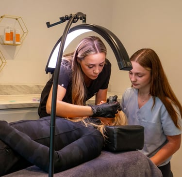 Profesional de microblading enseñando la técnica a alumna durante práctica en modelo con aro de luz en cabina blanca y beige.