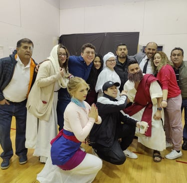 A diverse group of friends posing in a community hall wearing various costumes for a theatrical play or celebration.