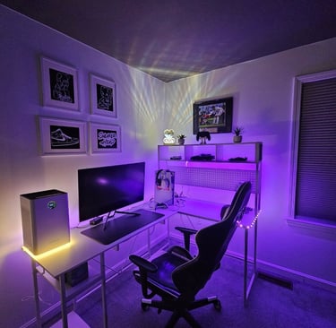 Modern gaming setup with an L-shaped desk, RGB lighting, and ergonomic chair in a purple-lit room.