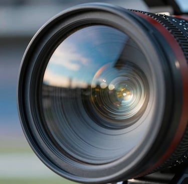 Macro photography of a professional cinema camera lens reflecting the bright LED lights of a North American / US sports arena. The reflections create patterns of sky blue and light gray-blue across the glass surface, embodying a sleek and high-tech aesthetic.